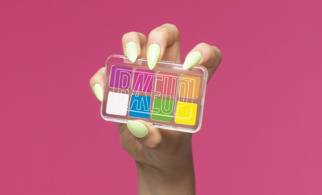 A promotional photo of the Bakeup Palette in Neon, in it, the palette is being held in a hand with green "claw" nails, on a bright pink background