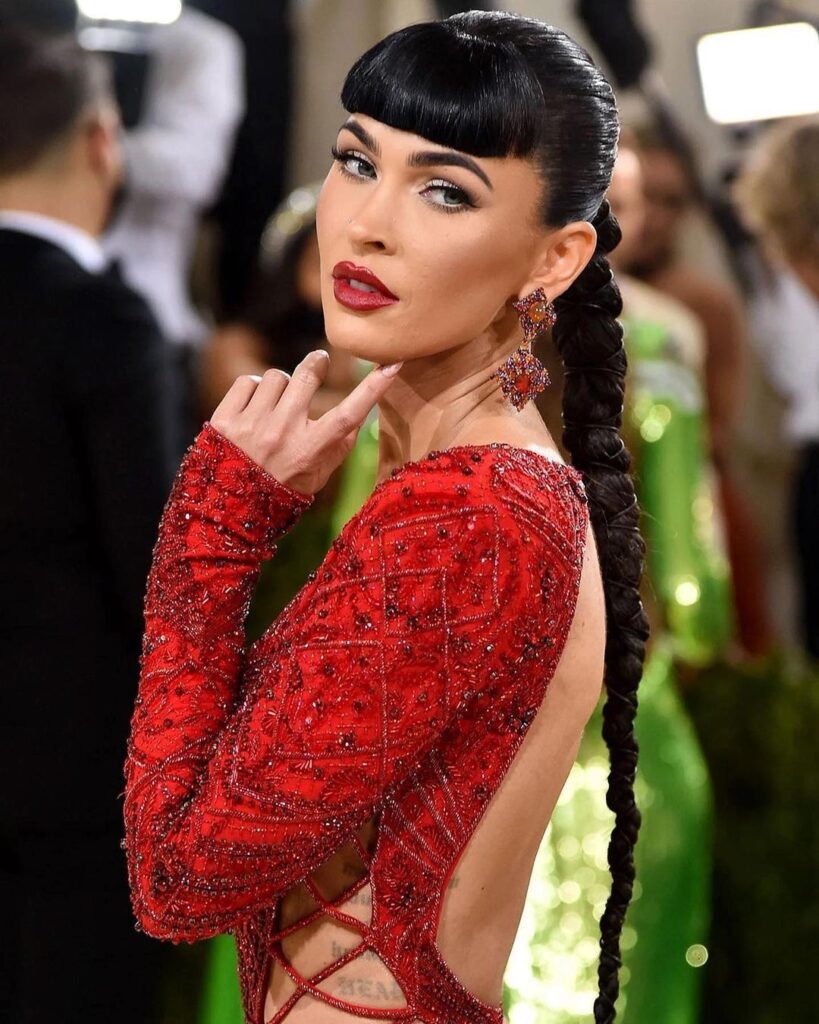A photo of Megan Fox, in which she is on the red carpet at the 2021 MET Gala, and is wearing a glittery red dress and has a long braid, and blunt bangs courtesy of Andrew Fitzsimons