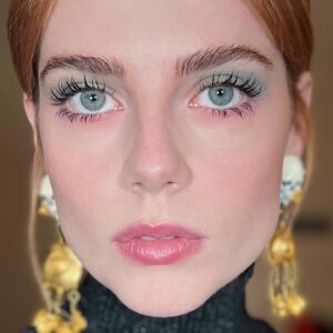 An upclose photo of Lucy Boynton where she is wearing dramatic blue and pink eyeshadow with long lashes and a pink lip, done by Jo Baker