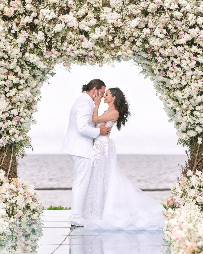 A photo of Scheana Shay and her husband Brock on their wedding day, in the photo they are both wearing white, in front of an arch made of pink and white flowers
